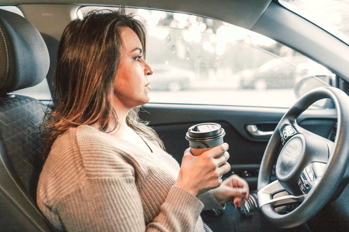 Une femme assise au volant de sa voiture à l'arrêt, un gobelet de café à la main, le regard tourné vers la route